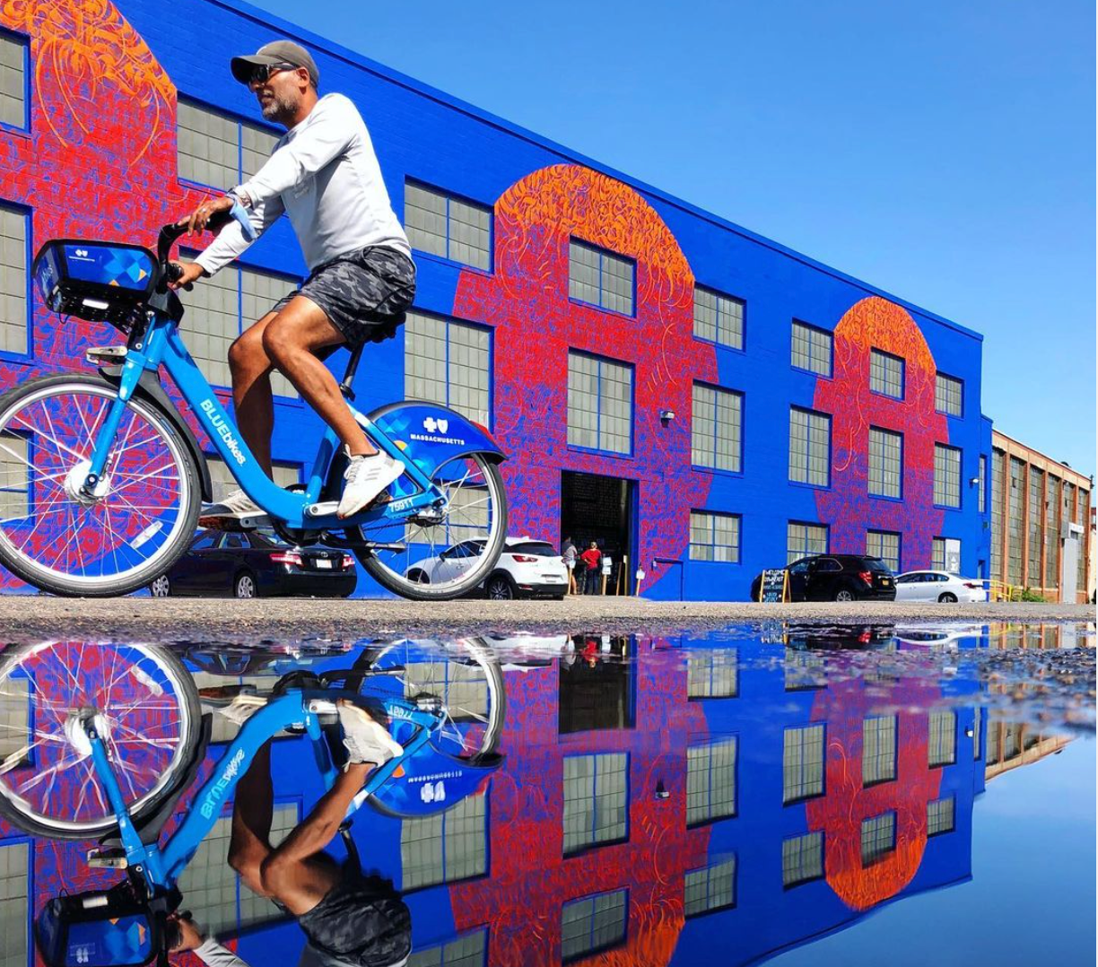 Pedestrians and bicycles passing a giant mural painted by IMAGINE (Sneha Shrestha) produced by HarborArts for Sea Walls Boston 2020. Photo by Christian Merfeld, Boston Harbor Now.